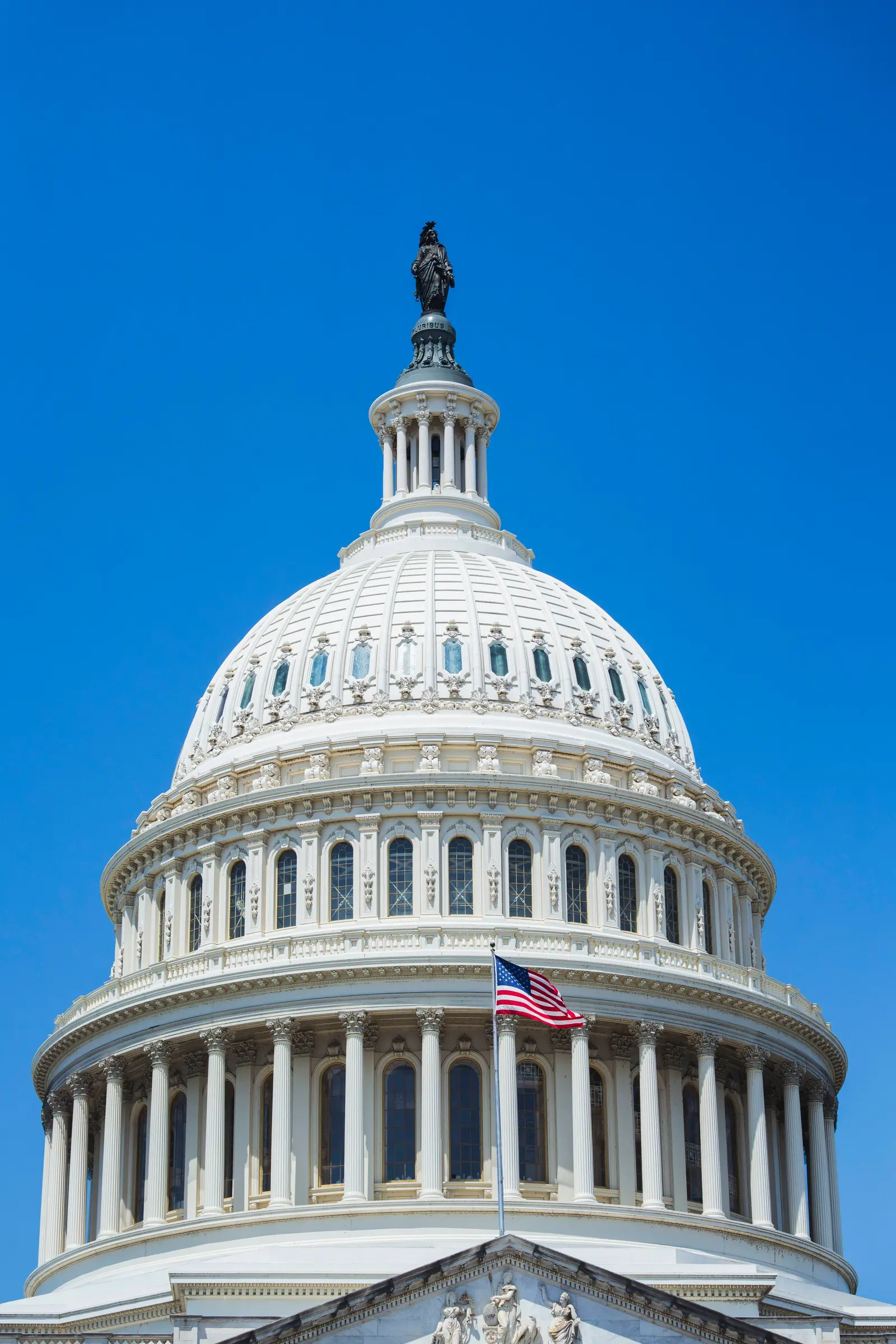 The United States Capitol dome with the flag flying, against a clear sky.